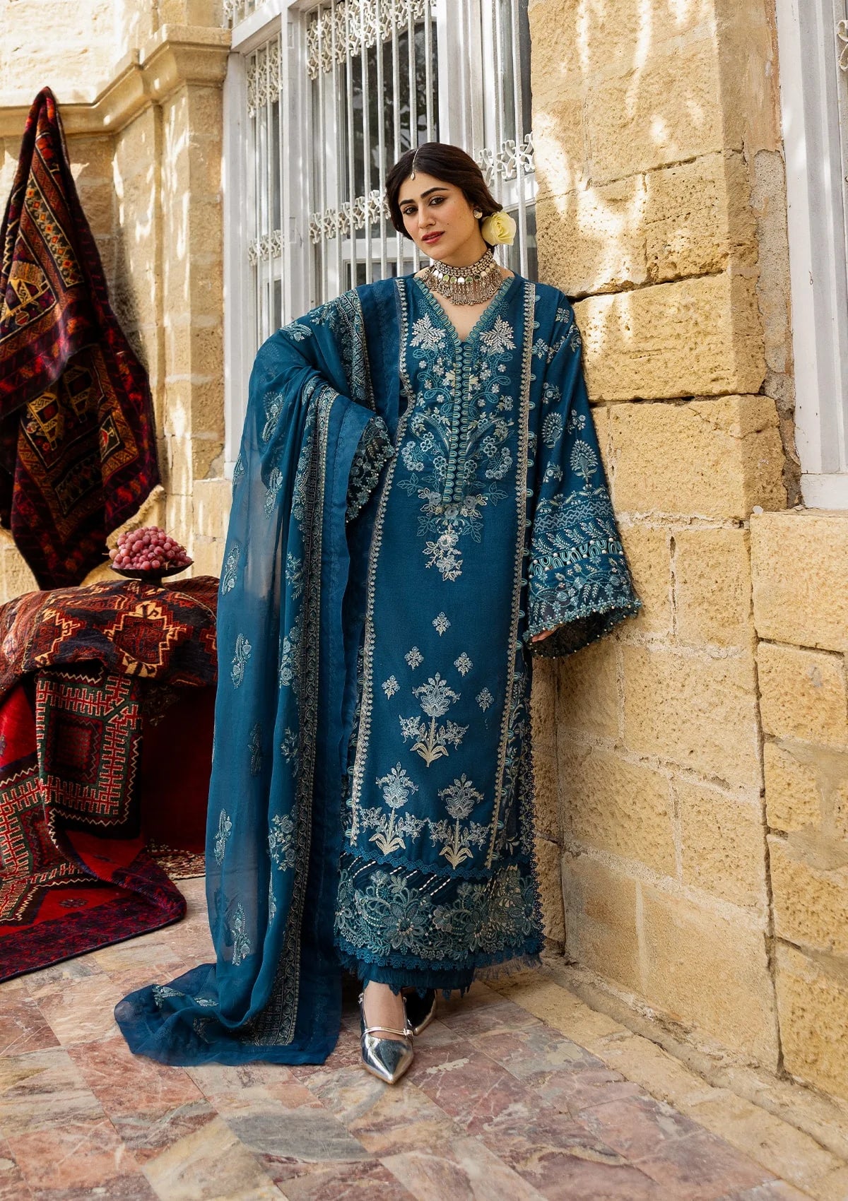 Woman in a blue embroidered traditional outfit standing in front of a stone wall.