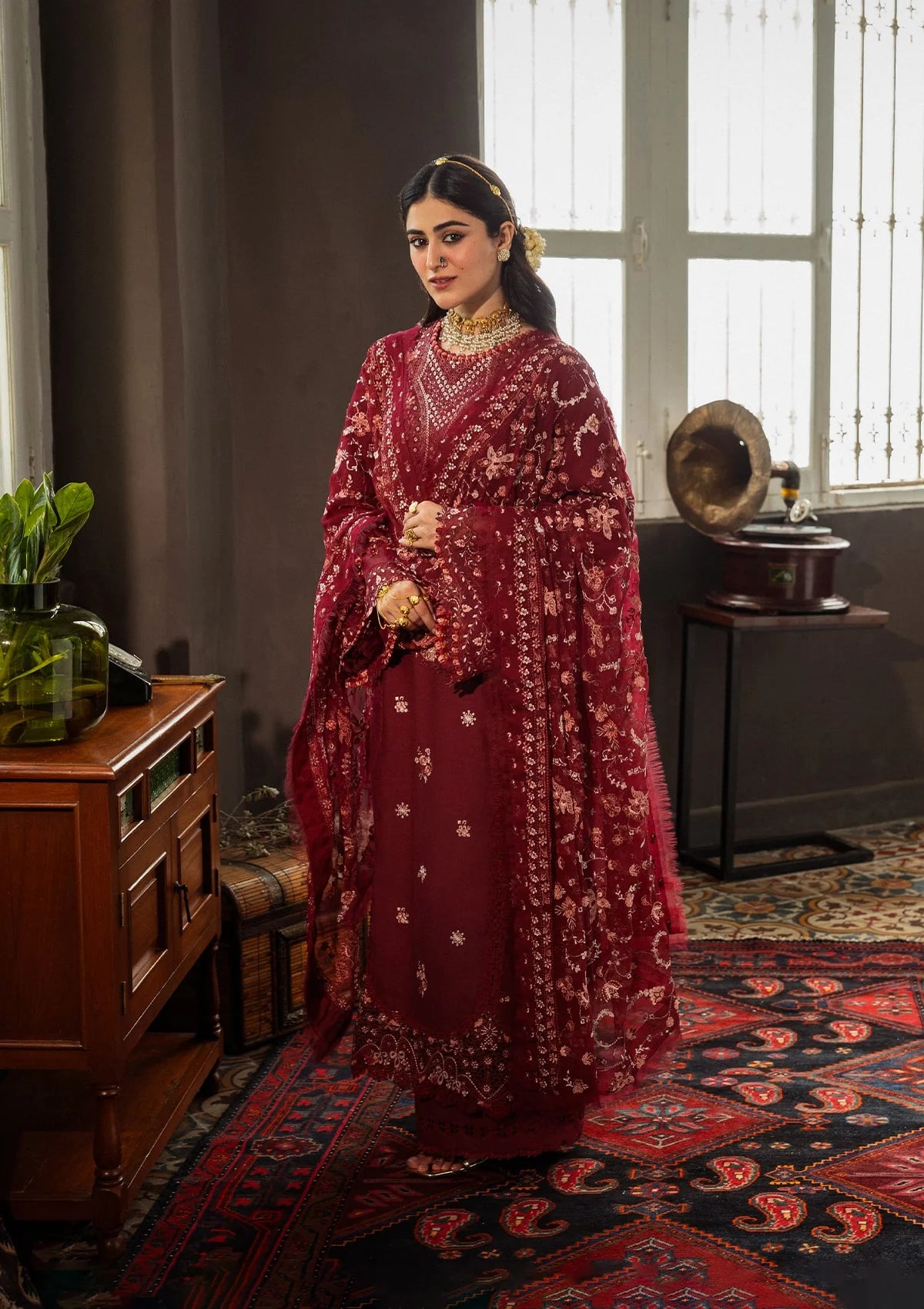 Woman in a red traditional outfit standing in a room with a window and wooden furniture.