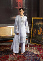 Woman in a light gray embroidered outfit standing in an ornate room with a painting and chair.