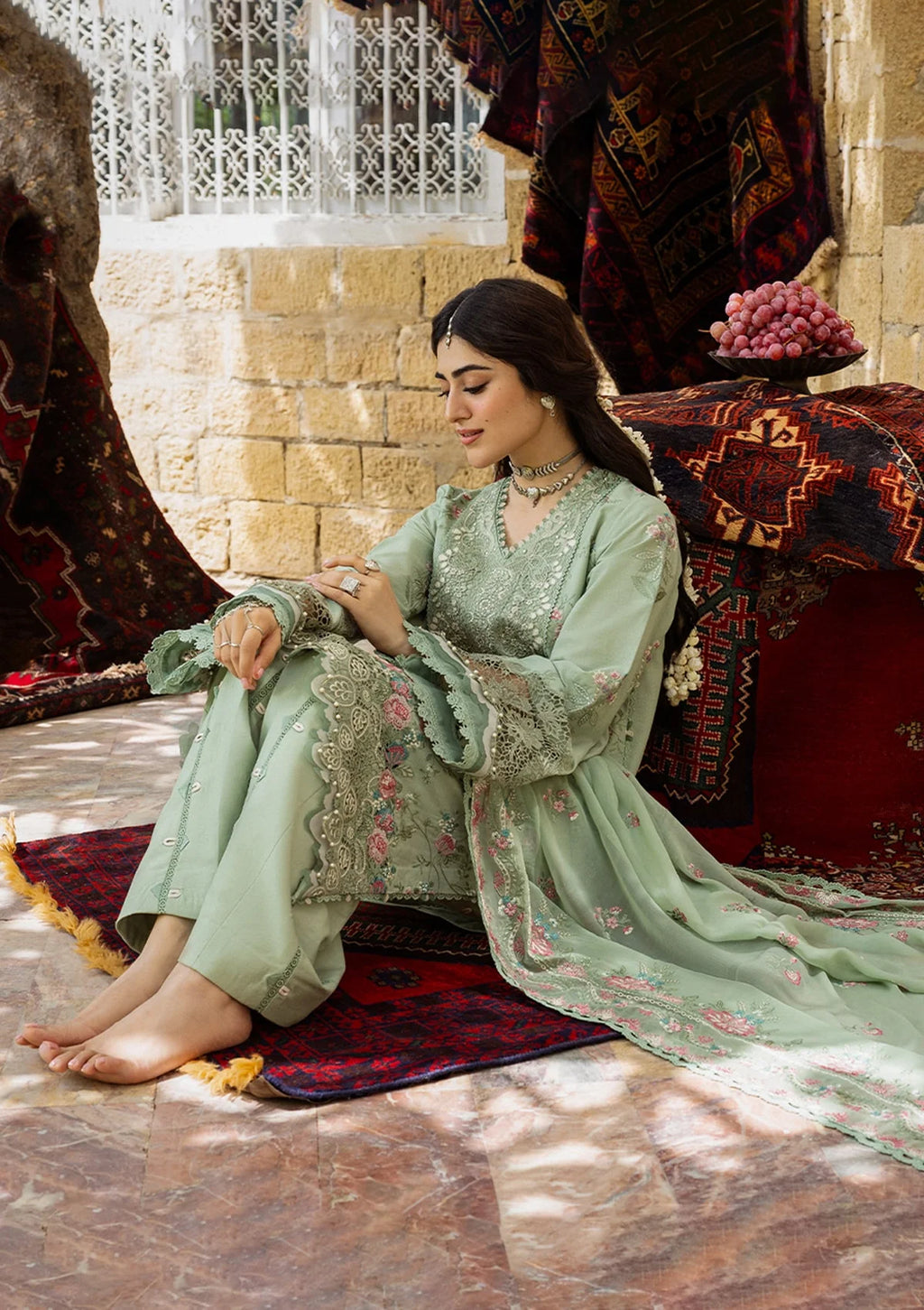 Woman in a light green traditional outfit sitting on a patterned rug with a decorative background.
