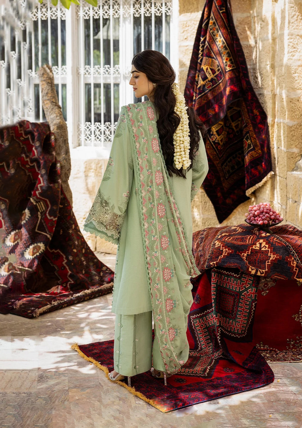 Woman in a light green traditional outfit with pink embroidery, standing in front of decorative rugs.