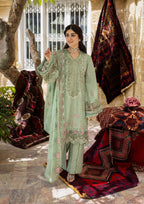 Woman in a light green traditional outfit standing in front of decorative rugs and a tree.