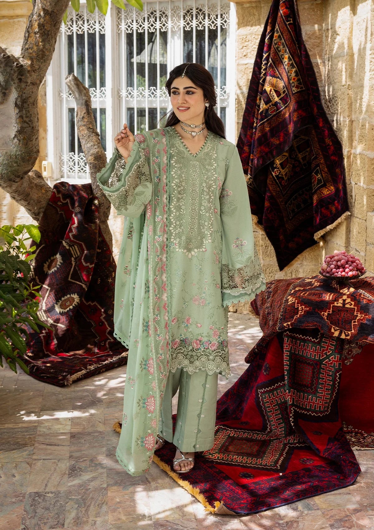 Woman in a light green traditional outfit standing in front of decorative rugs and a tree.